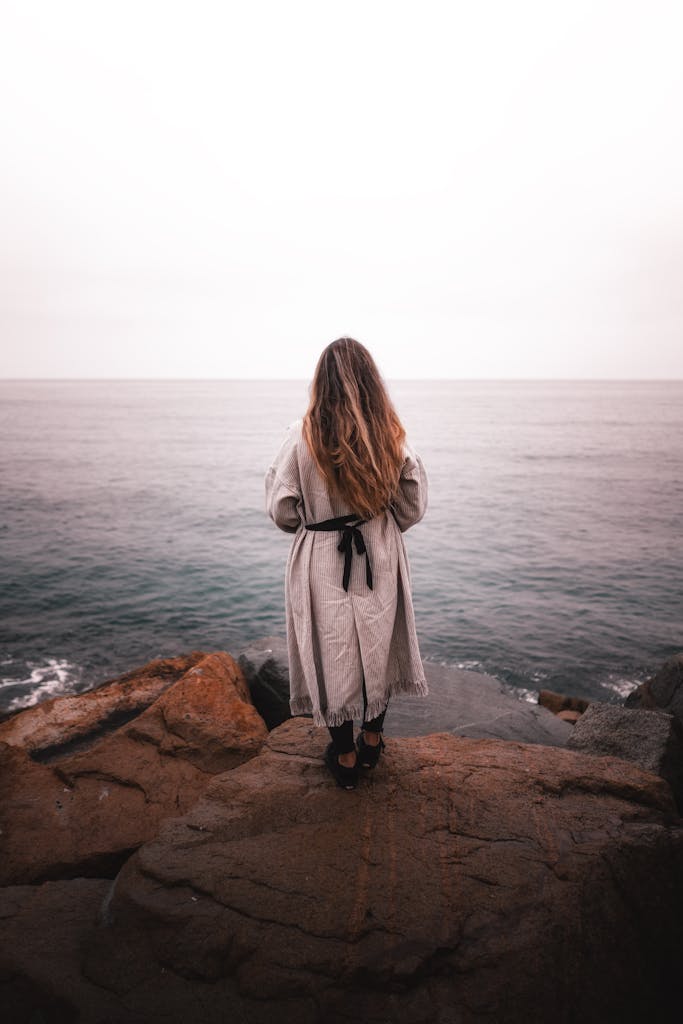 A woman stands on a rocky shore, contemplating the calm sea in solitude.