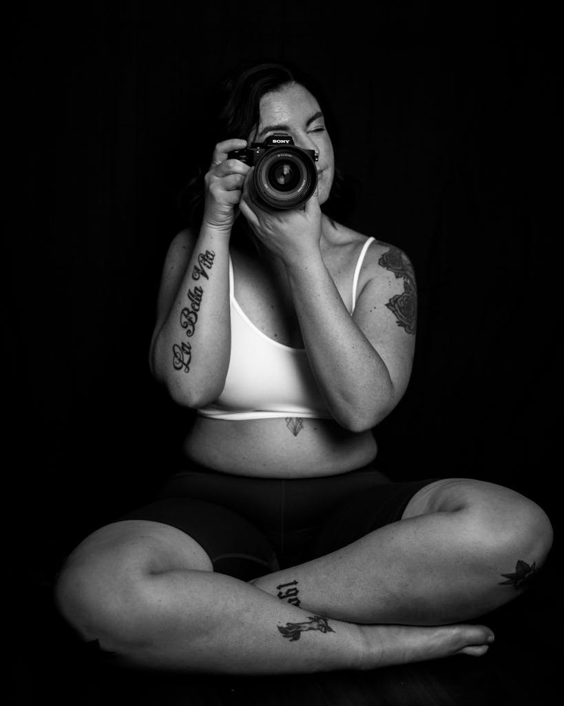 Plus-size tattooed woman sitting cross-legged, holding a camera in a B&W artistic pose.