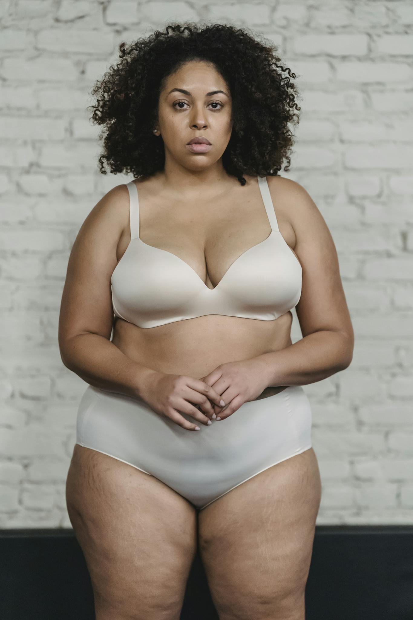 Portrait of a confident plus-size woman in lingerie against a brick wall, promoting body positivity.