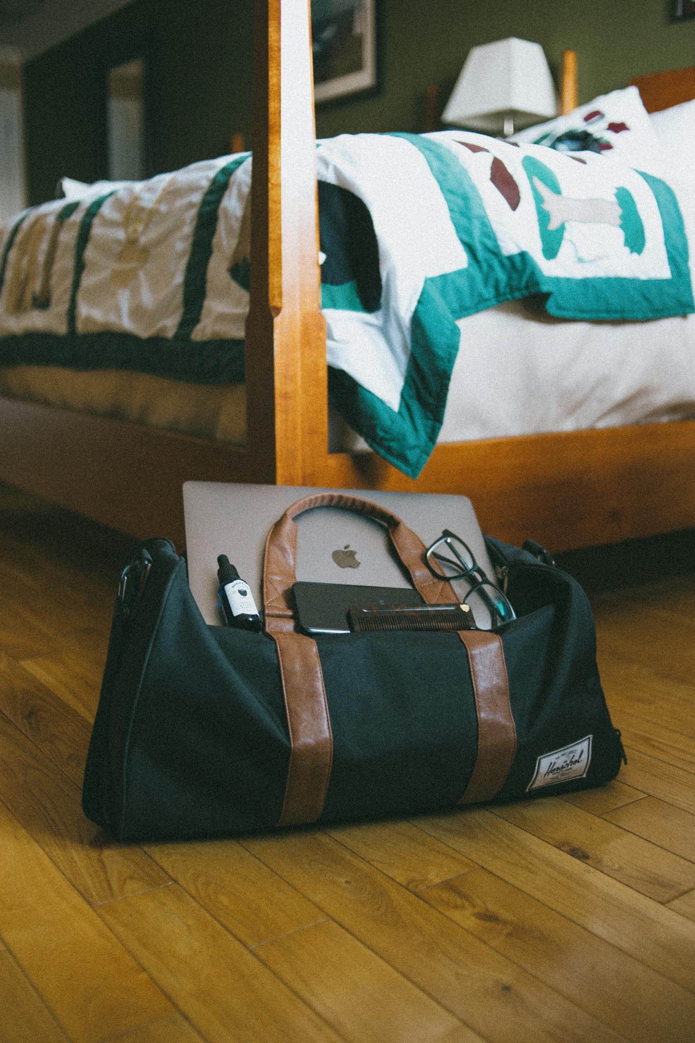 Travel bag on wooden floor with gadgets near a cozy bed, highlights travel readiness.