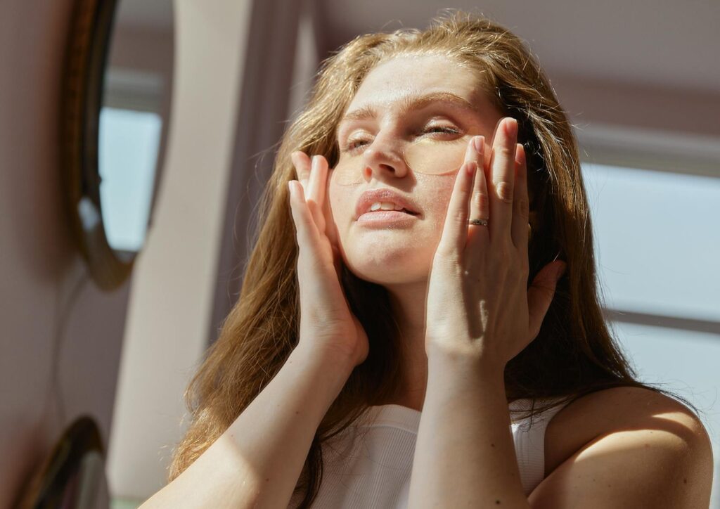 Woman in white tank top applies skincare products while enjoying the sunlight in her home.