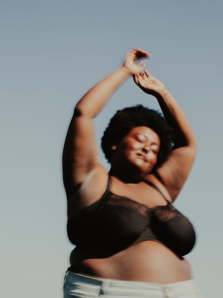 Joyful woman in a bra celebrates body positivity outdoors under a clear sky.