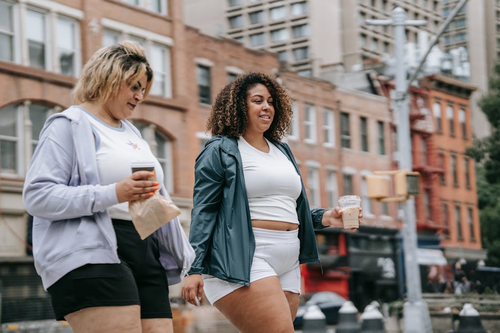 Two women walking in the city, enjoying drinks and laughter on a warm day.