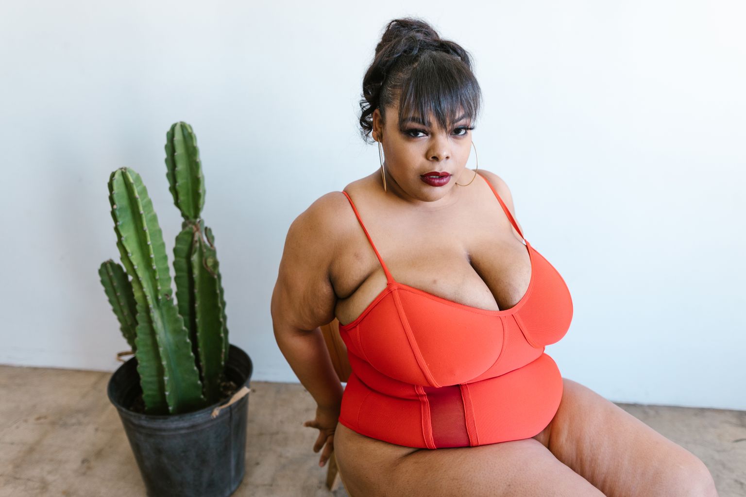 A body-positive woman in a red swimsuit poses beside a cactus indoors.