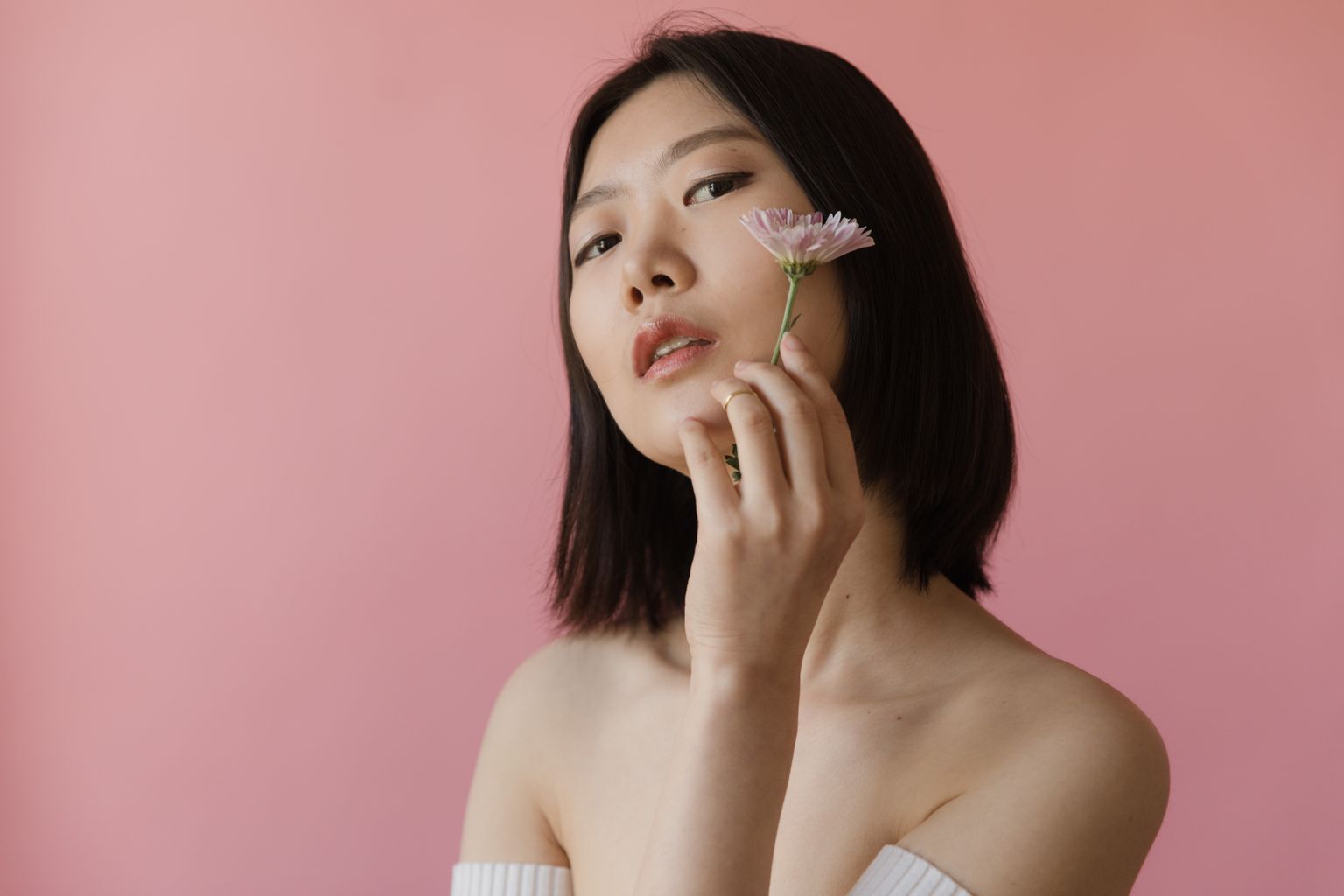 A close-up portrait of an Asian woman with short hair holding a pink flower against a pastel pink background.