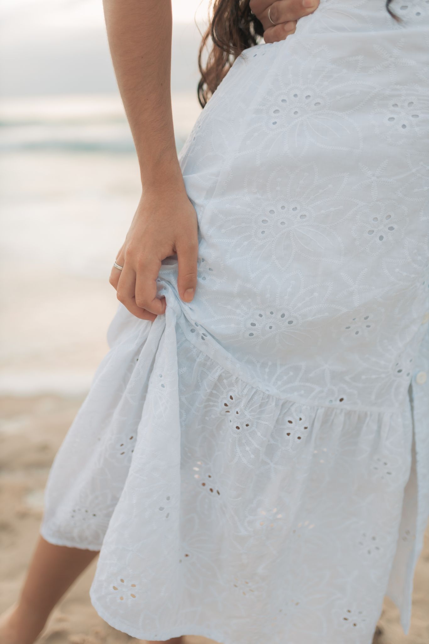 A partial view of a woman gently holding her white eyelet dress at the beach.