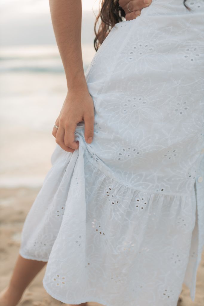 A partial view of a woman gently holding her white eyelet dress at the beach.