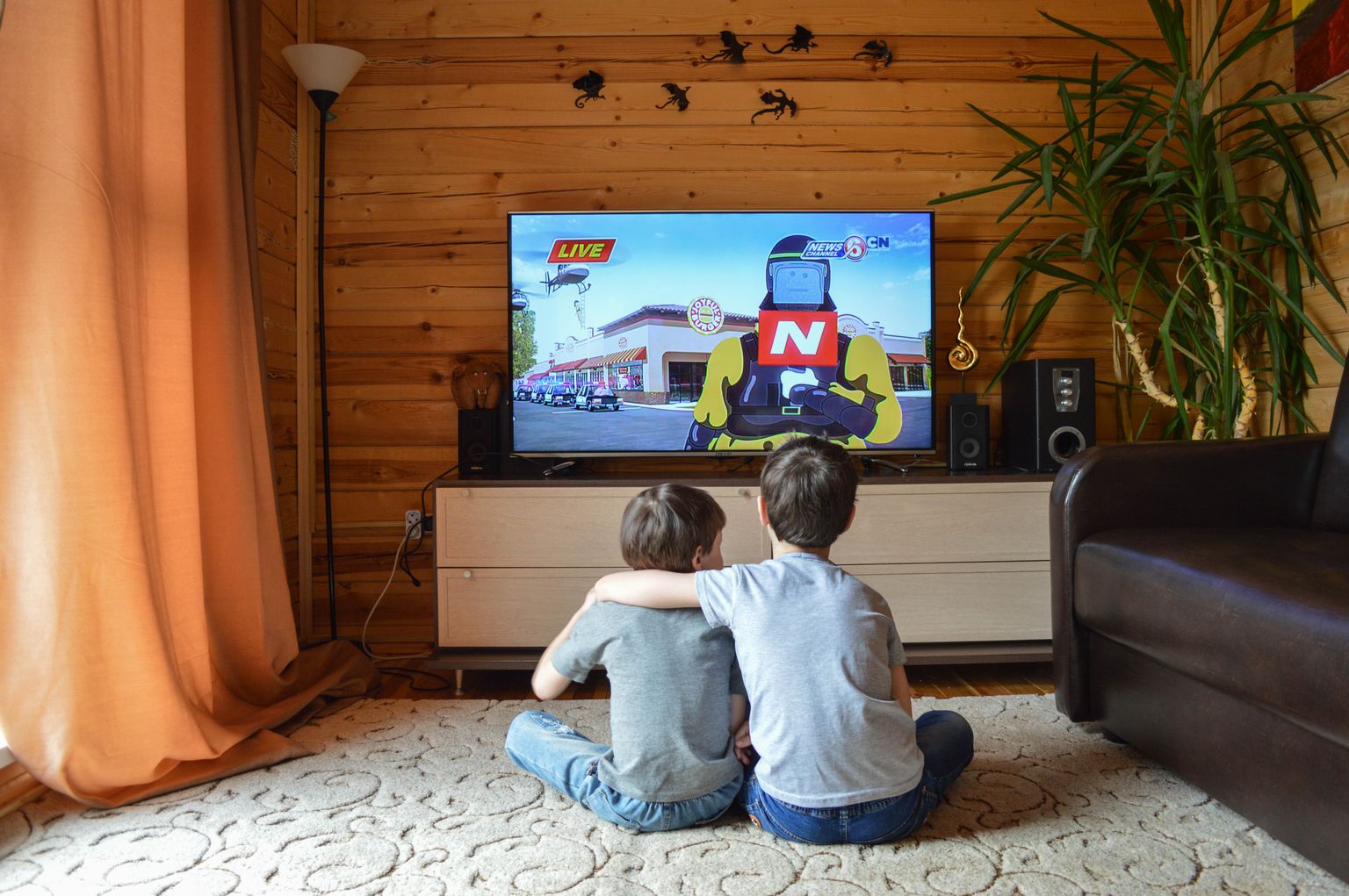 Back view of unrecognizable little children in similar casual outfits resting on floor at home and hugging while watching cartoon on TV