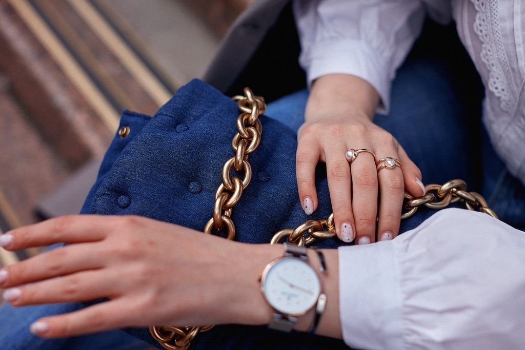 Close-up of fashionable accessories including a blue handbag with a gold chain and jewelry on hands.