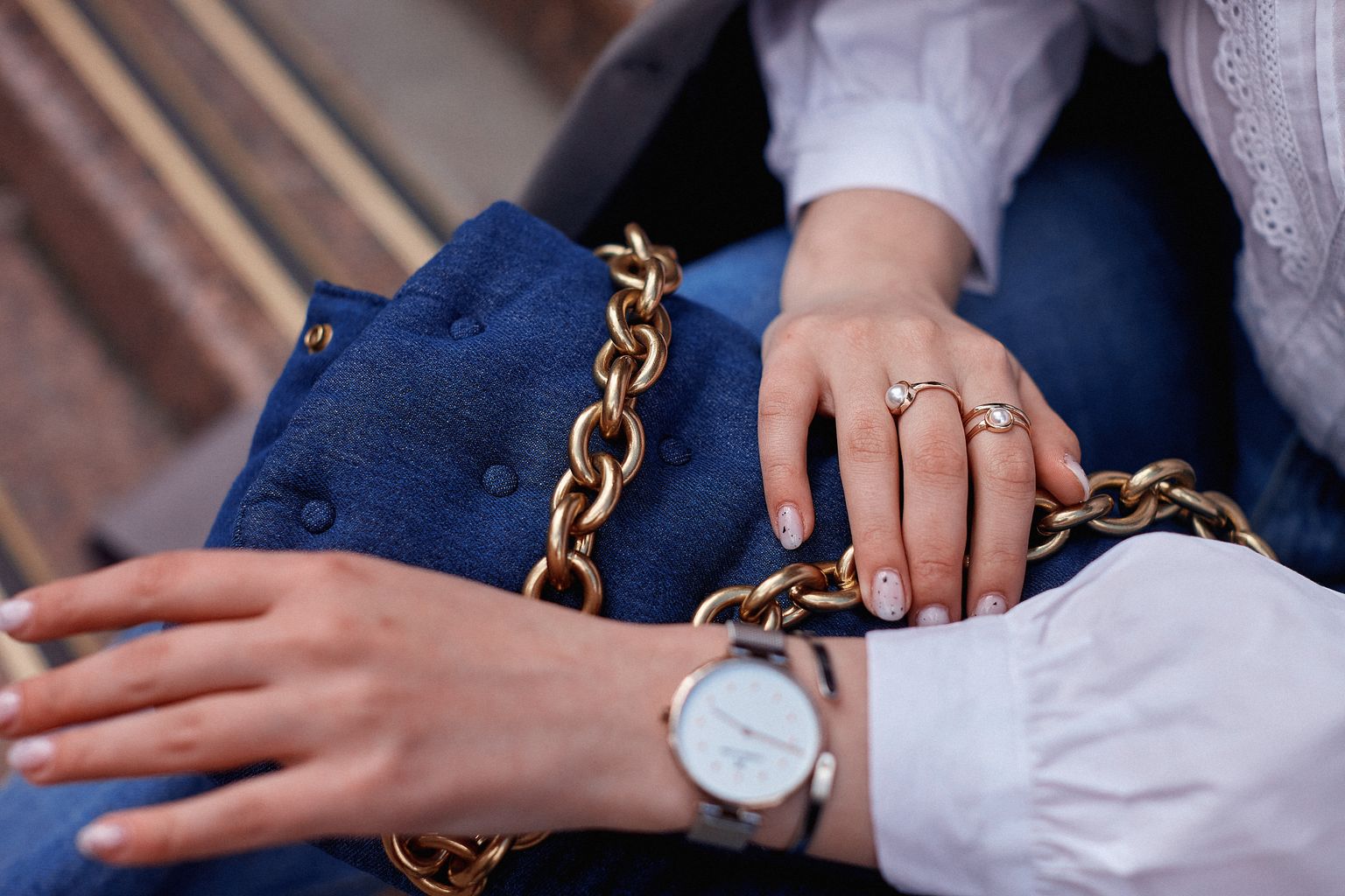 Close-up of fashionable accessories including a blue handbag with a gold chain and jewelry on hands.