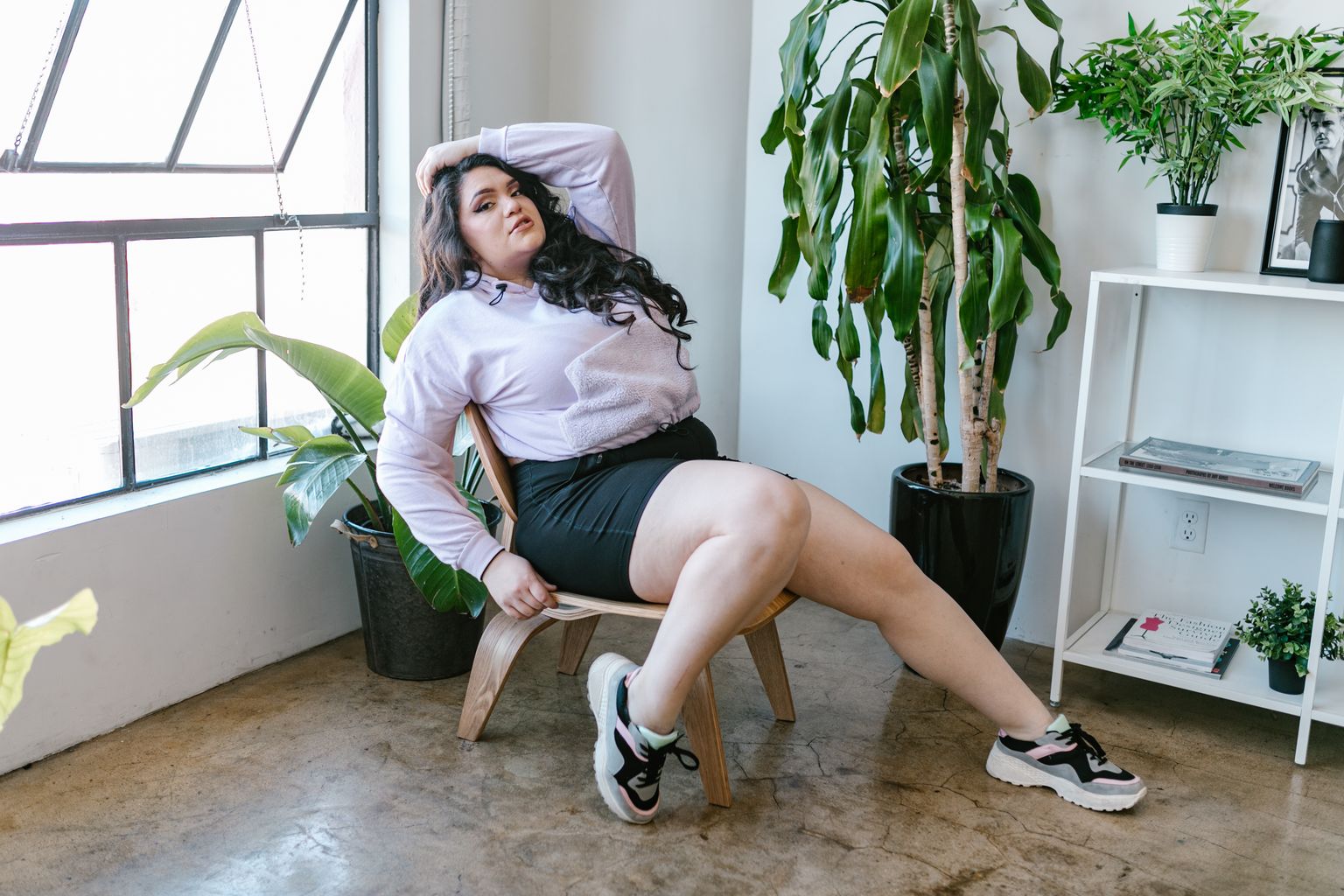 Confident woman posing on a chair with lush green plants in a bright room.