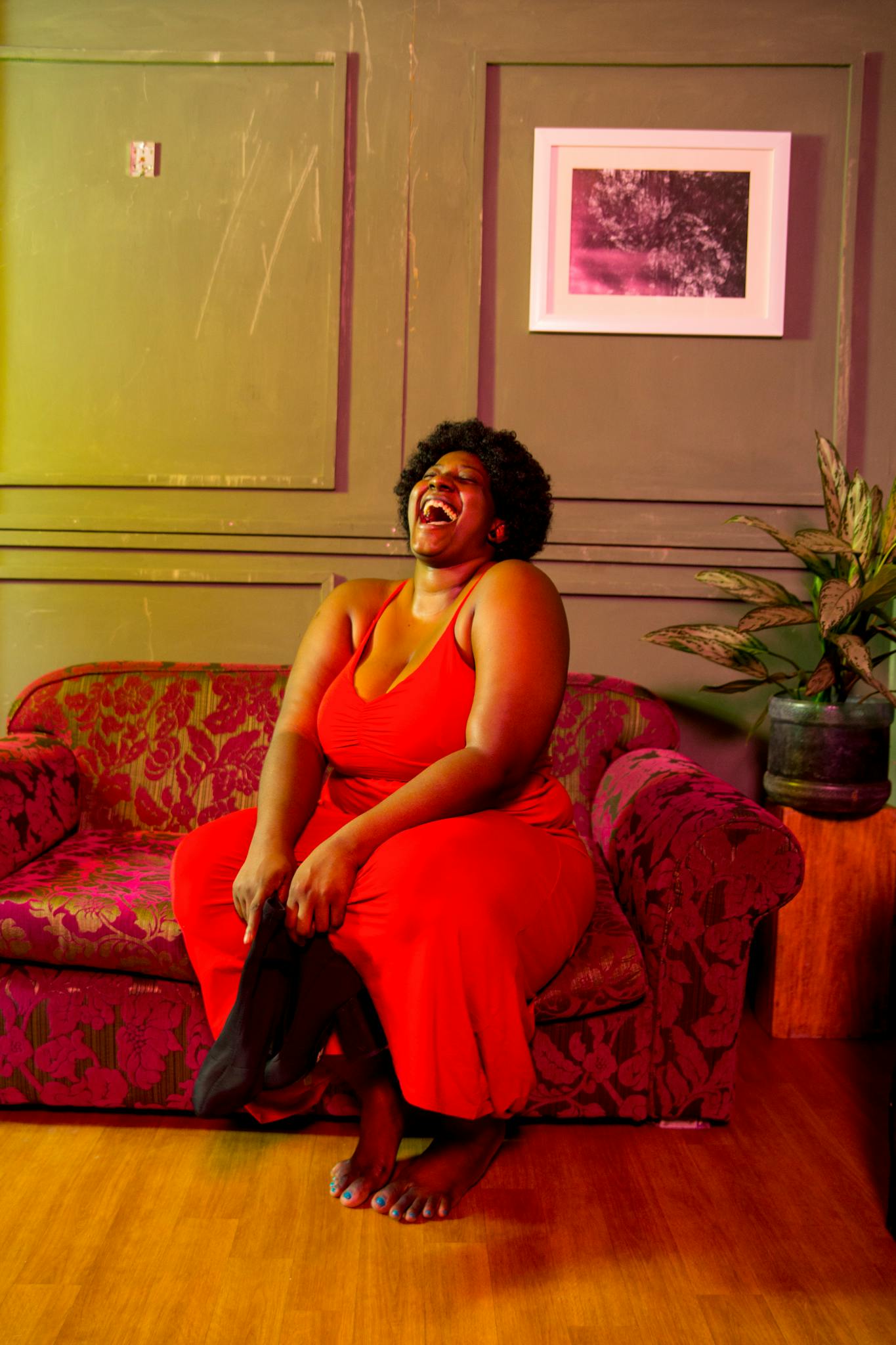 Full body of barefoot plump African American female laughing with mouth opened sitting on comfortable sofa