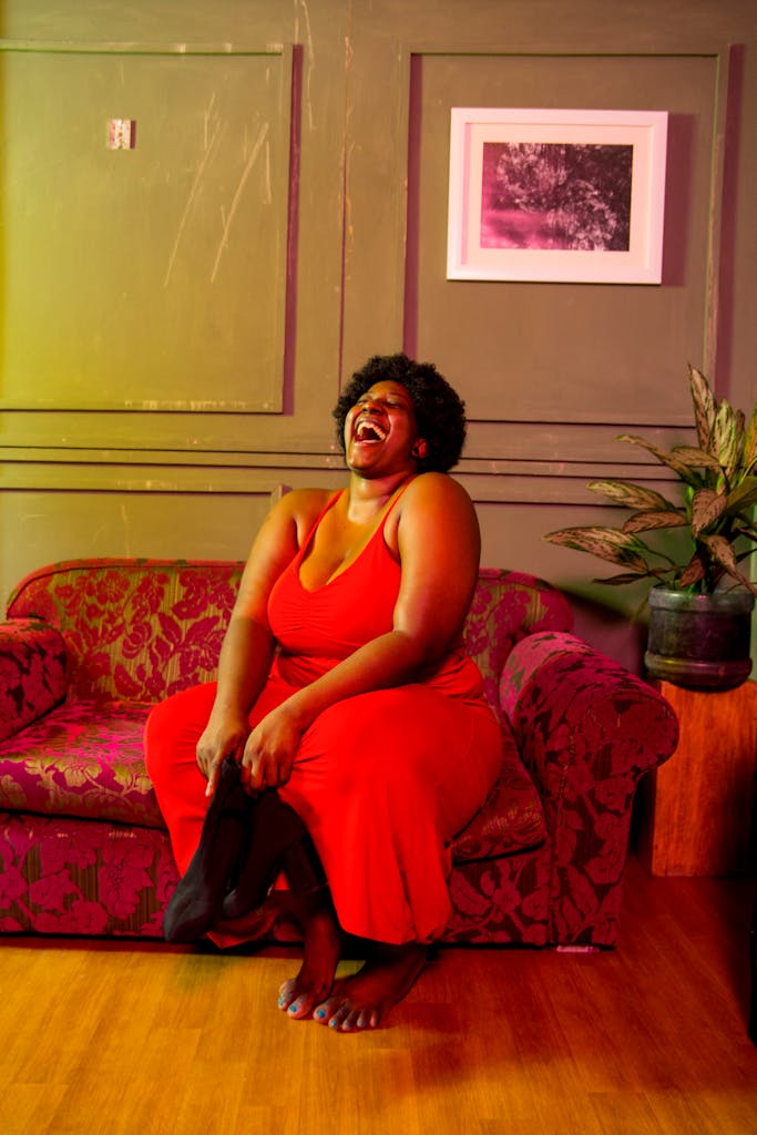 Full body of barefoot plump African American female laughing with mouth opened sitting on comfortable sofa