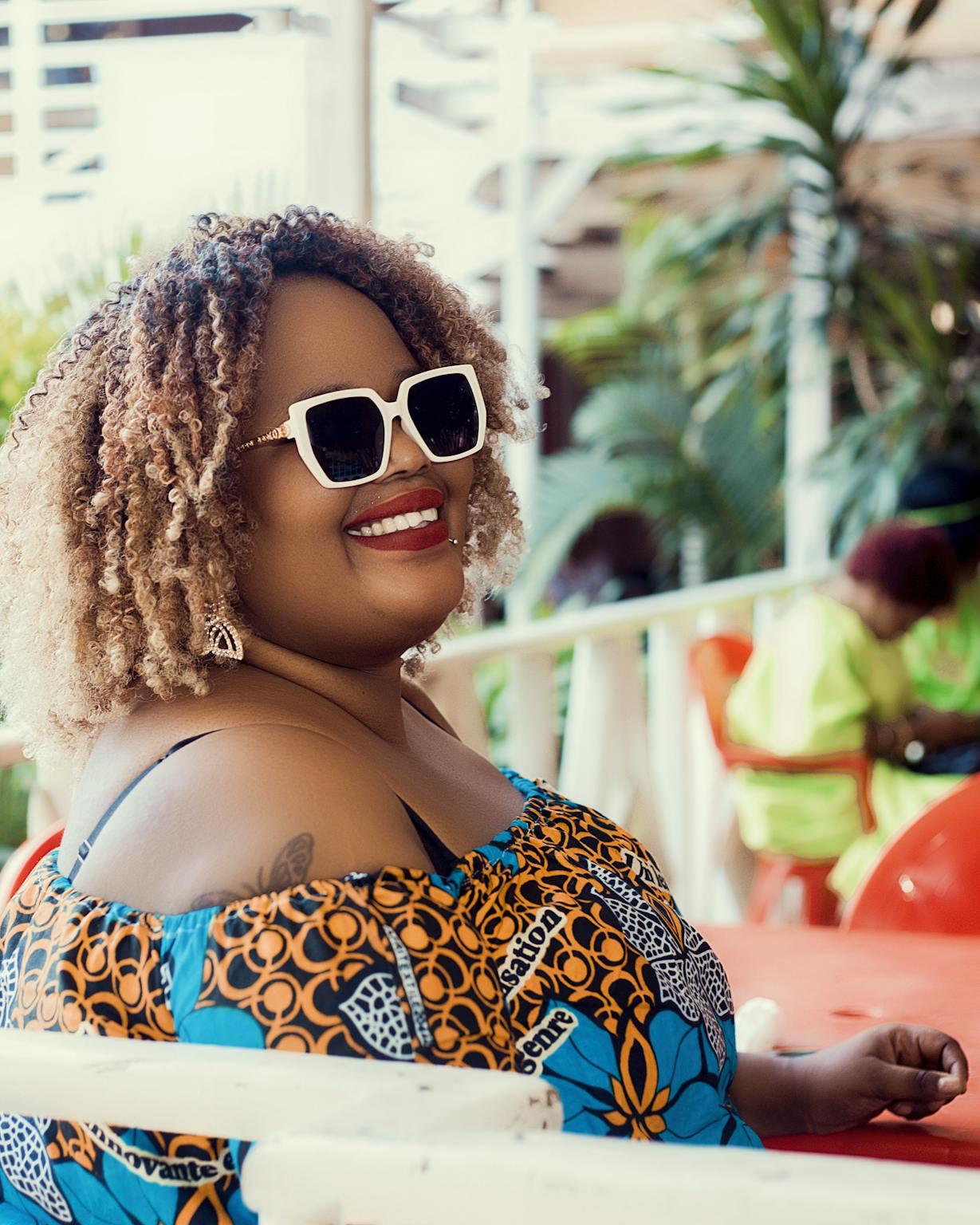 Joyful woman wearing sunglasses and colorful dress, enjoying a sunny day at an outdoor café.