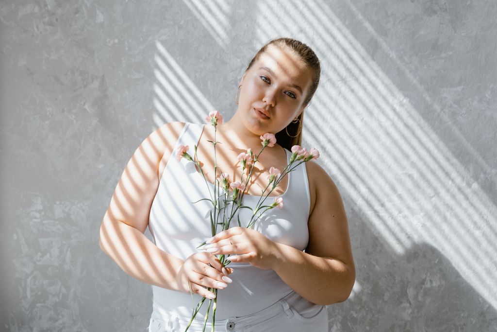 plus size woman in tank top holding flowers with sunlight filtering through blinds. 8433431