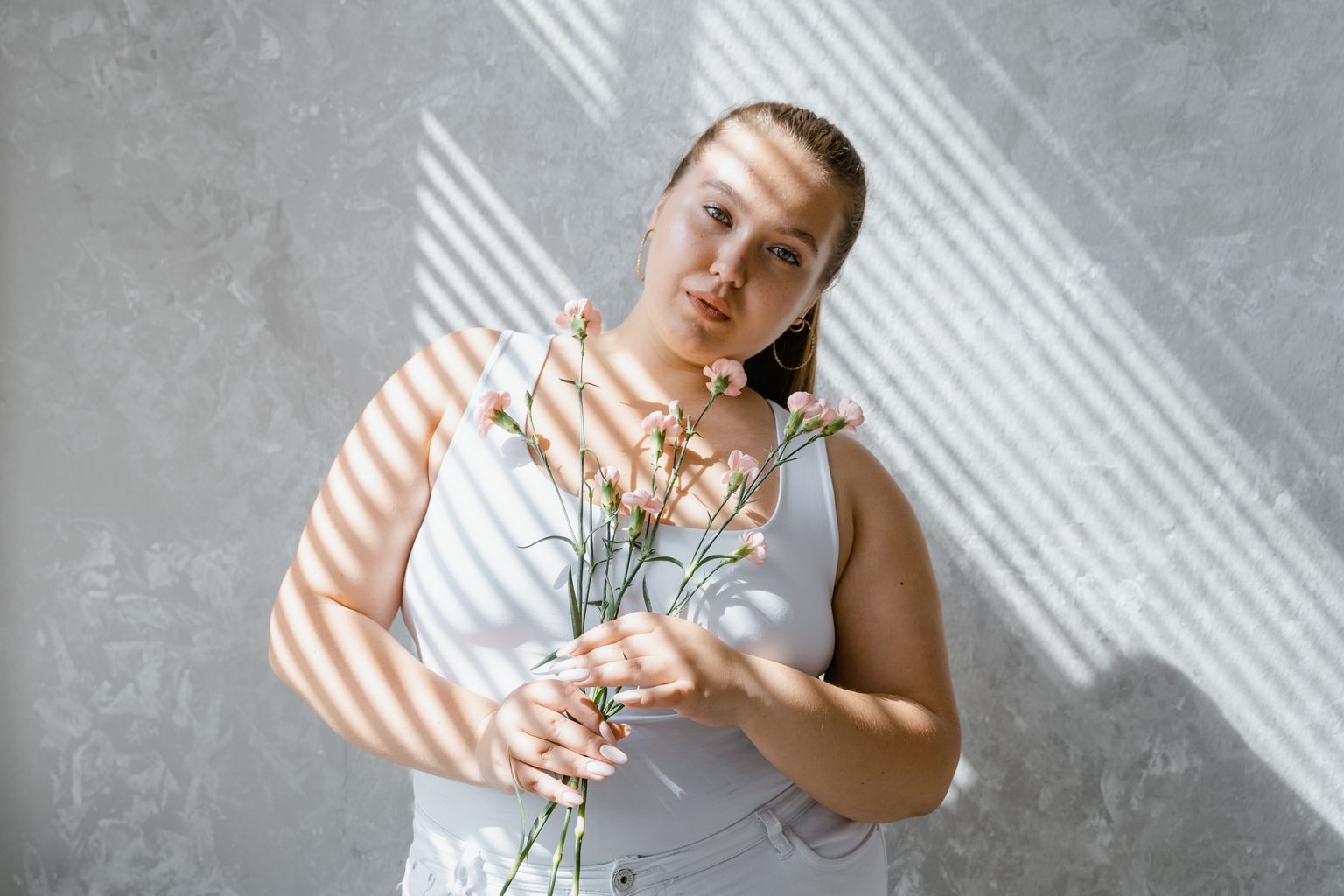 plus size woman in tank top holding flowers with sunlight filtering through blinds. 8433431