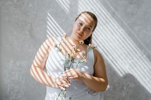 plus size woman in tank top holding flowers with sunlight filtering through blinds. 8433431