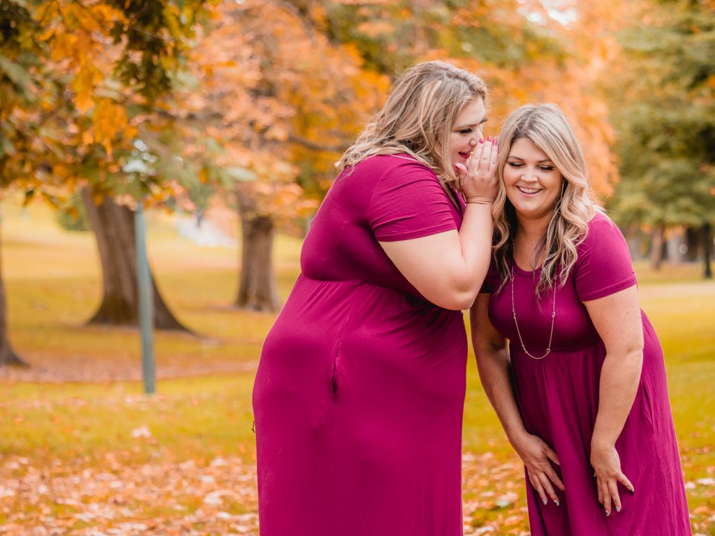 Two happy women in pink dresses gossip in a vibrant autumn park setting.