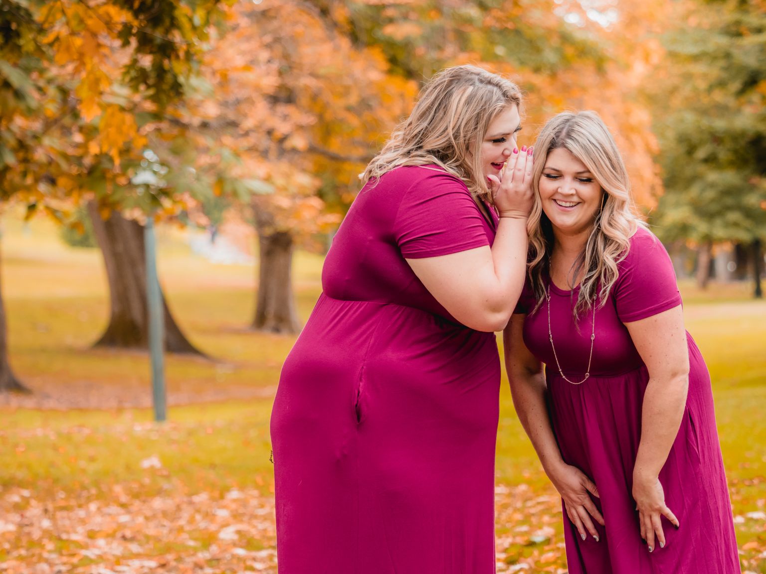 Two happy women in pink dresses gossip in a vibrant autumn park setting.