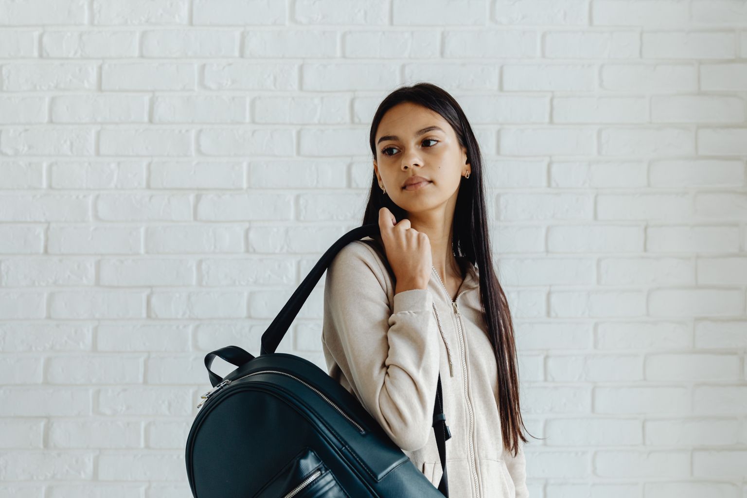 Young woman in casual attire with backpack, standing indoors against a white brick wall.