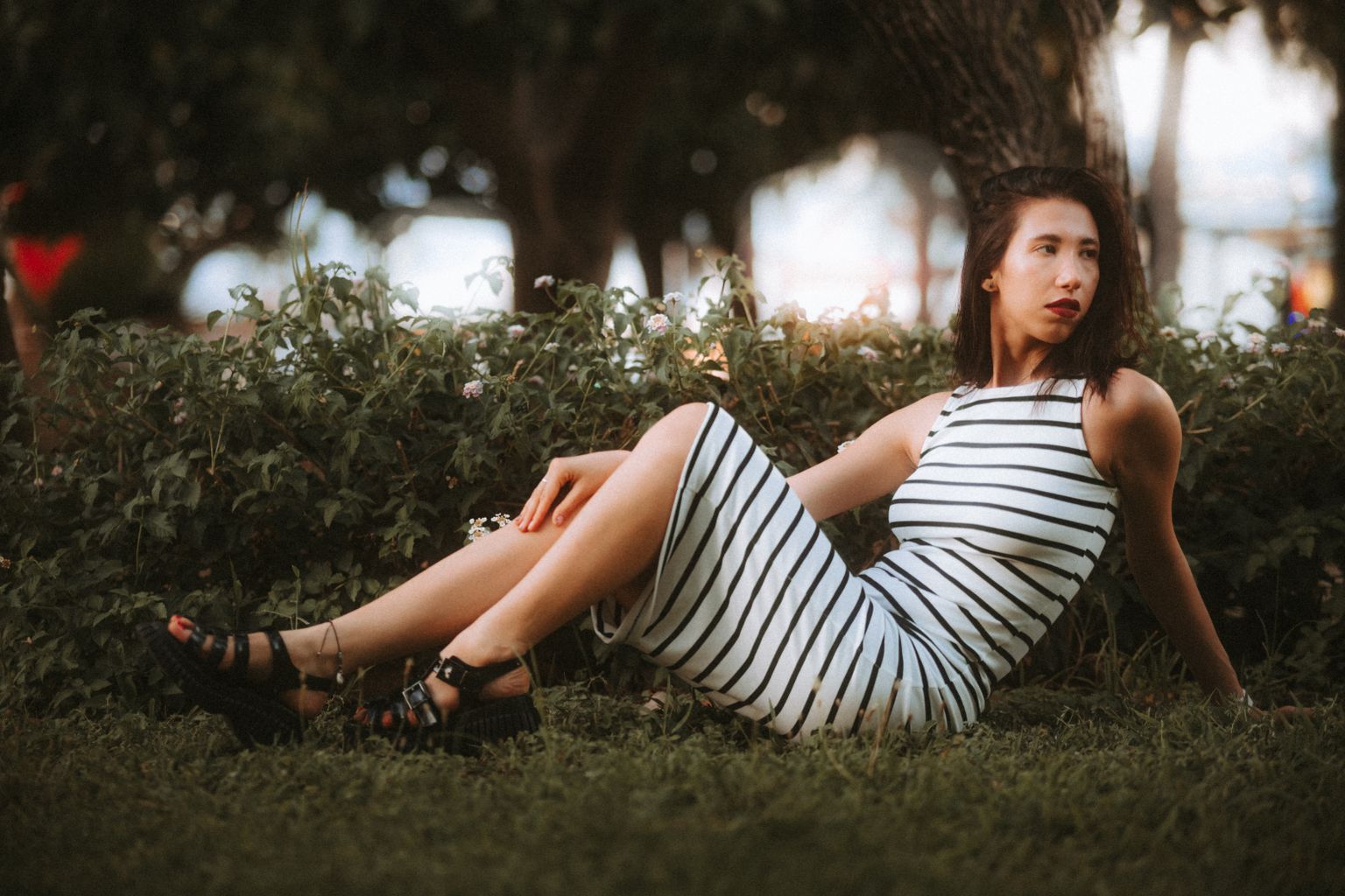Fashion-forward woman in a striped dress sitting gracefully in a lush park setting.