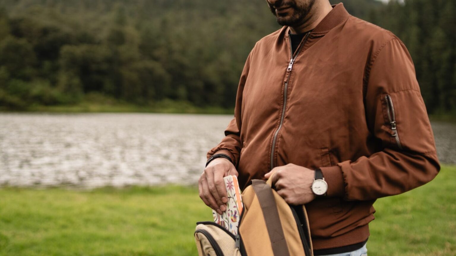Man placing item in backpack by a scenic lakeside, wearing a casual brown jacket.