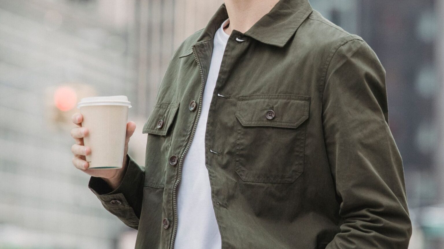 Side view of crop faceless young male in trendy outfit standing on modern city street with paper cup of coffee