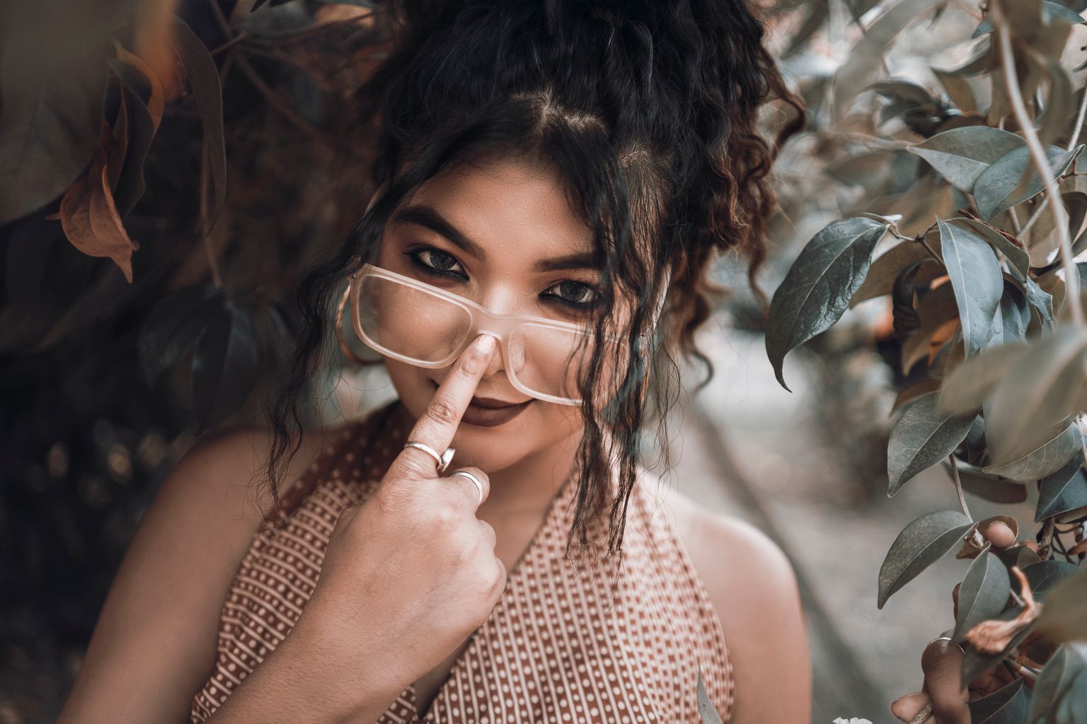 South Asian woman playfully adjusting glasses amidst leafy surroundings.