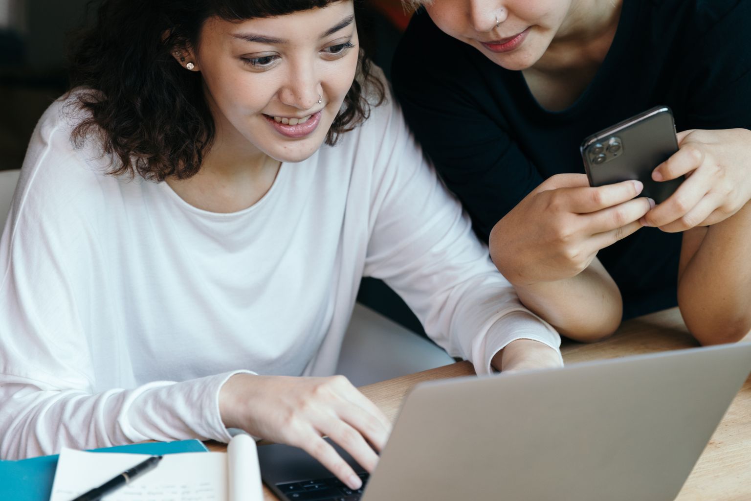 Young girlfriends in casual clothes working together on laptop and using mobile phone while developing collaborative project