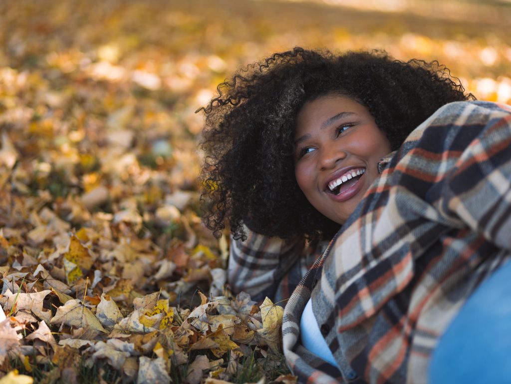 A cheerful woman with curly hair enjoying a relaxing moment lying on autumn leaves outdoors.