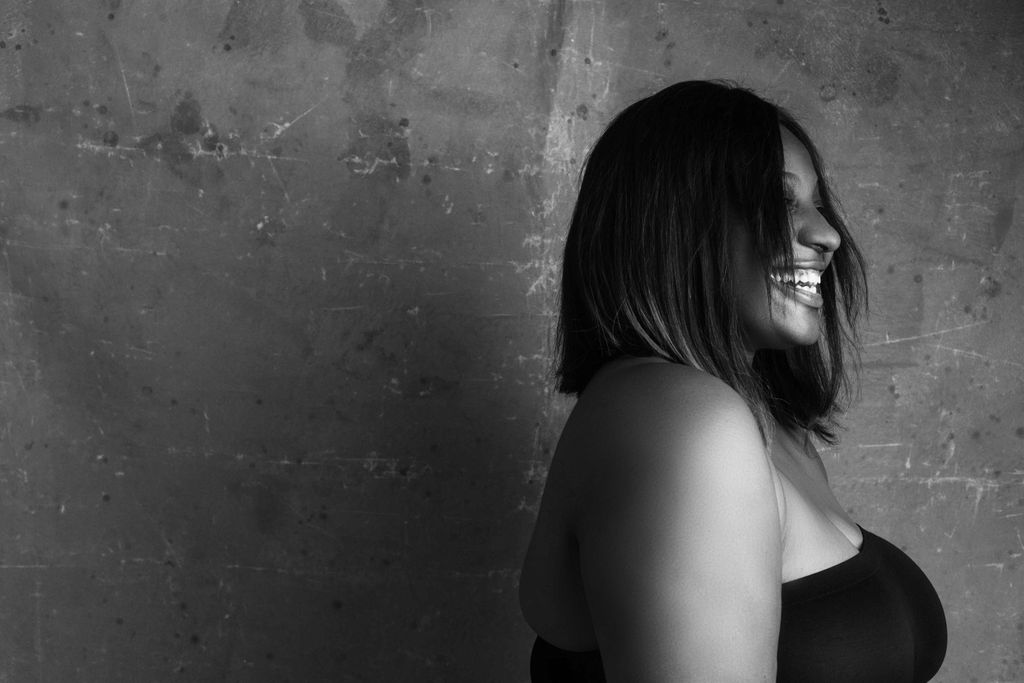 Black and white side view of African American plus size female laughing happily while standing against grungy gray wall