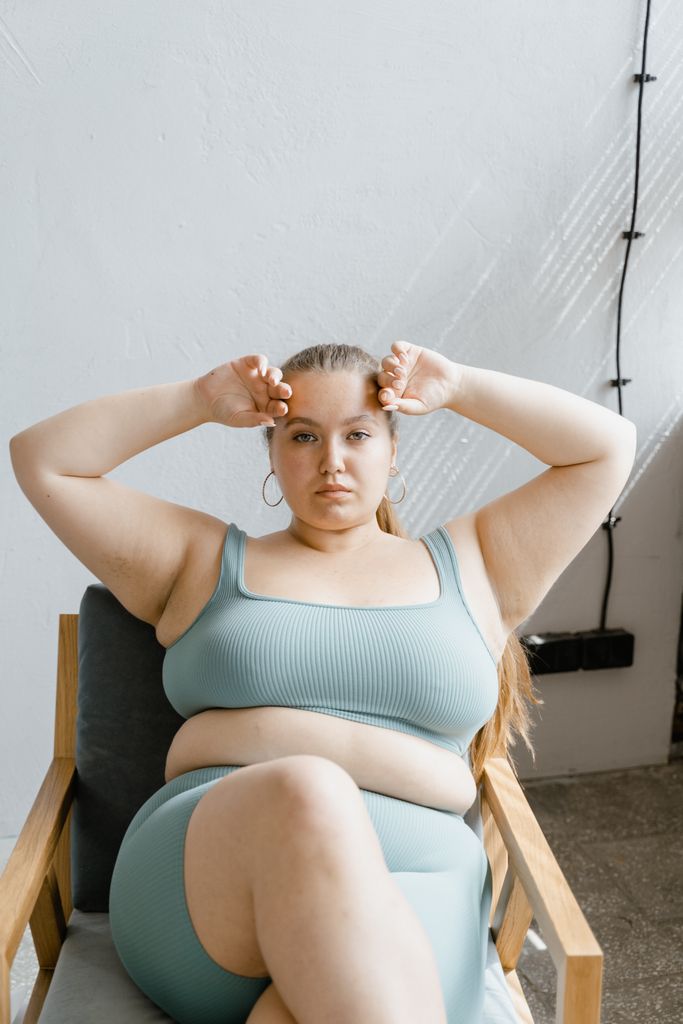 Portrait of a confident woman sitting in a chair indoors, wearing a teal bralette, with hands on head.