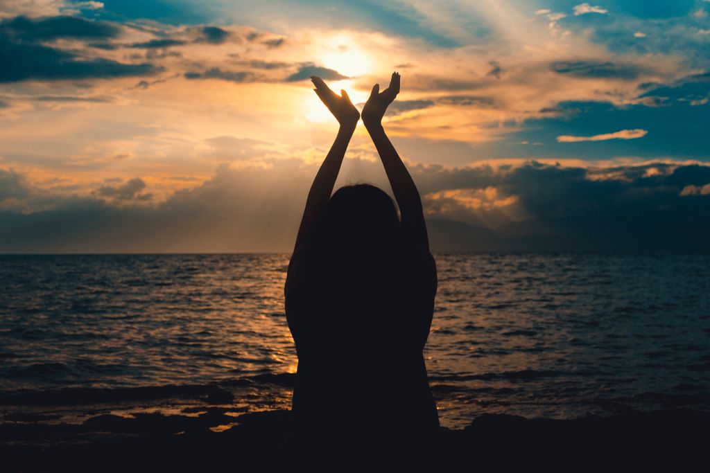 A silhouetted figure raises hands towards the setting sun over a serene ocean view.